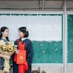 Two graduates celebrate in front of a bulletin board with a sunflower bouquet and diploma.
