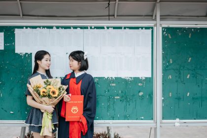 Two graduates celebrate in front of a bulletin board with a sunflower bouquet and diploma.