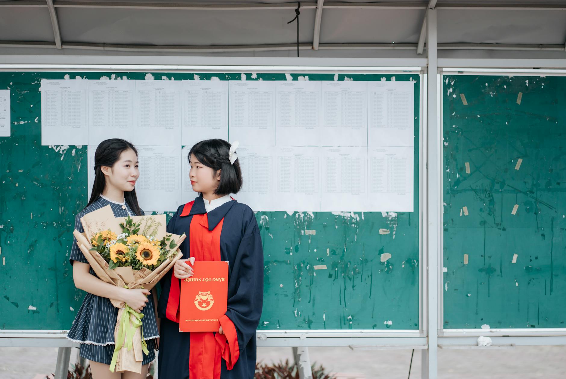Two graduates celebrate in front of a bulletin board with a sunflower bouquet and diploma.