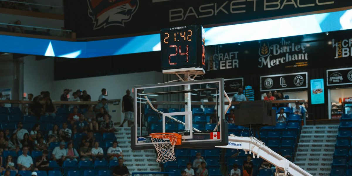 Close-up of a basketball hoop inside a stadium with a scoreboard and audience in the background.